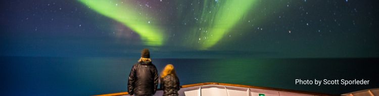 A couple stands on the tip of a boat looking at auroras.